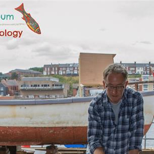 Artist Stephen Livingstone works on a sculpture and there are boats, houses and a river in the background. 