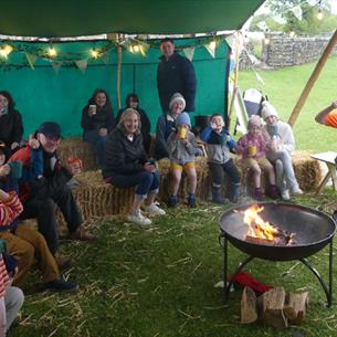 Families and children relax on hay bales under a green canopy tent, holding cups of hot drinks and gathered around a small fire pit, with open country