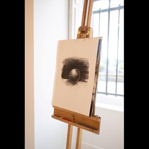 A wooden easel holding a drawing pad with a charcoal sketch of a sphere against a dark shaded background. Natural light from a window behind. 