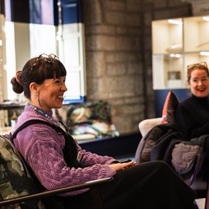 Two people seated indoors in a cozy, well-lit room with stone and blue walls, and large windows. One person wears a purple sweater and s
