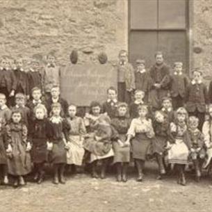 An old fashioned school photograph of children sat in lines in front of a school building 