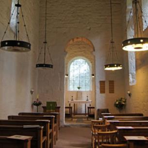 The interior of an Anglo Saxon church, with white stone walls and hanging round lights.