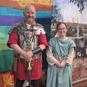 Two people dressed in Roman clothes, smiling in front of information boards