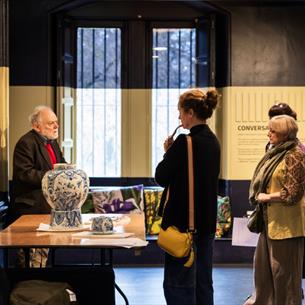 Photograph of a museum or gallery setting where a guide explains exhibits to a group of four visitors. The scene features display cases with artifacts
