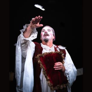 A person on a dark stage wearing dramatic white face pain in historical robes, reaching out for a feather.