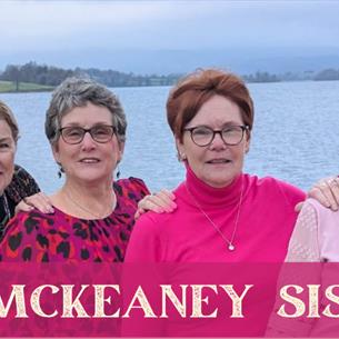Four sisters, smiling shoulder to shoulder in pink clothing, in front of a body of water. The text 'The McKeaney Sisters' underneath.