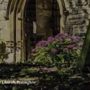 An arched stone church doorway, surrounded by purple flowers in the sunlight.