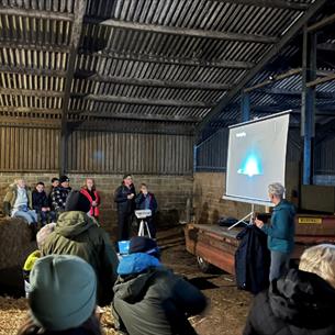 A group of people sit on hay bales inside a barn while a speaker gives an astronomy presentation beside a projected image.