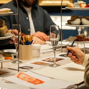 Photograph captures a table table during a shared meal, with small plates and diners eating, drawing and selecting food from tiered serving stands. Pl