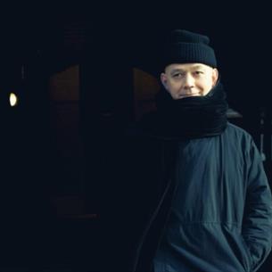 A person wearing dark clothes, a hat and scarf, standing smiling in a dark hallway.