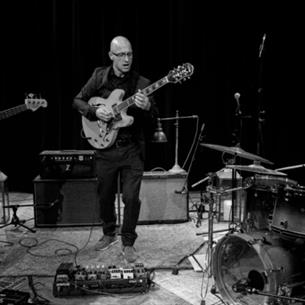 Three people playing their respective instruments, bass, guitar and drums in a rehearsal space. The image is in black and white.
