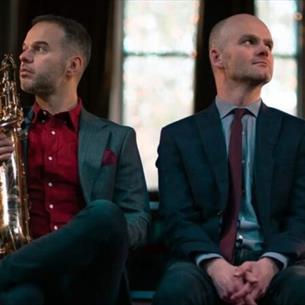 Two people sat facing away from each other, wearing suits and holding a brass instrument.