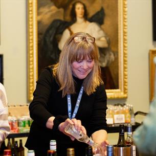 Photograph of women conducting a food or drink tasting at a museum event, with bottles, samples and name badges arranged on a table. A large framed po
