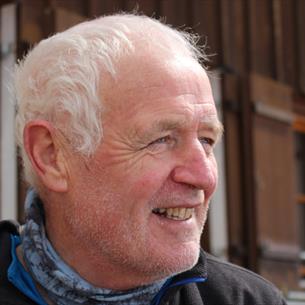 Photograph showing an older white-haired man outdoors beside a timber-clad building. He smiles in profile, wearing a black jacket and blue-grey neck w