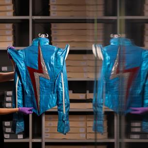 Photograph showing a curator in purple gloves displaying a bright blue metallic costume inside a clothing archive. Shelves of labelled storage boxes a