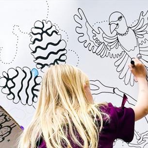 Photograph shows a child in a purple top drawing on a illustrated wall while sitting on an adult's shoulders. Wall features black line-art birds, fish