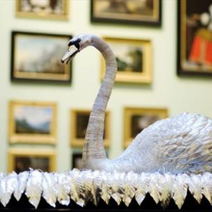 Photograph of a silver swan displayed in a gallery or museum, surrounded by framed artworks. Curved neck, textured feathers and ornate leafy base form
