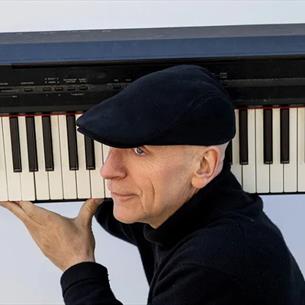 Person wearing a black flat cap and a turtle neck, carrying a piano keyboard over their shoulder against a grey background