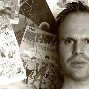 Alex Collier looking quizzical, shoulders up, in front of a wall of vintage comic books, the image is in black and white.