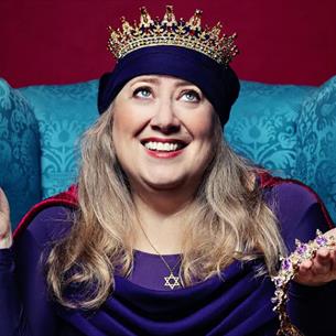 Person wearing a crown, sat on a blue chair holding two crown other crowns on a burgundy background.