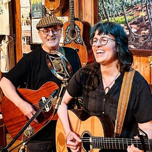 Two people playing guitars, smiling in a folk-like venue