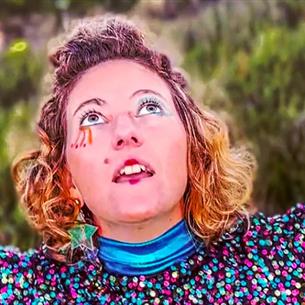 Person wearing colourful sequins and makeup, looking up bewildered with arms open, in front of nature.
