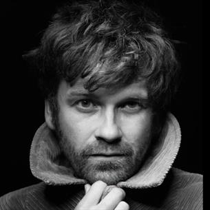 Black and white photograph of a man with tousled hair and a beard, looking directly at the camera. 