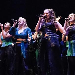 Group of people on a dark stage, singing wearing sparkly blue outfits