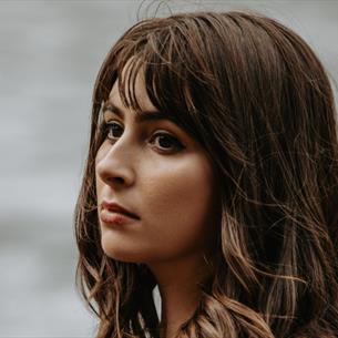 Photograph of a woman with shoulder-length brown hair, wearing a black top, looking pensively to her left. Background features a softly blurred body o