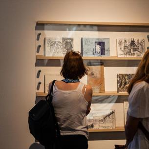 People observing framed sketches and drawings displayed on a gallery wall, highlighting an art exhibition setting. 