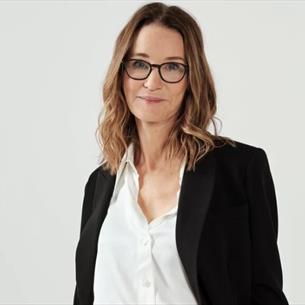 Susie Dent in a black blazer against a grey background.