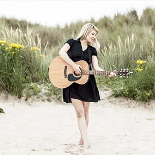 Hattie Murdoch playing the guitar barefoot on the beach, grassy dunes with yellow flowers make up the background.