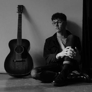 Black and white photo of John Gatenby sitting against a wall with his knees drawn up, guitar beside him.