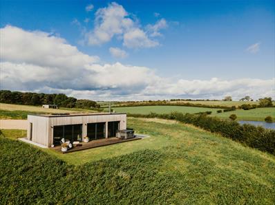Modern wooden lodge with large glass windows and hot tub set in a grassy hilltop overlooking green fields and a pond under a blue sky