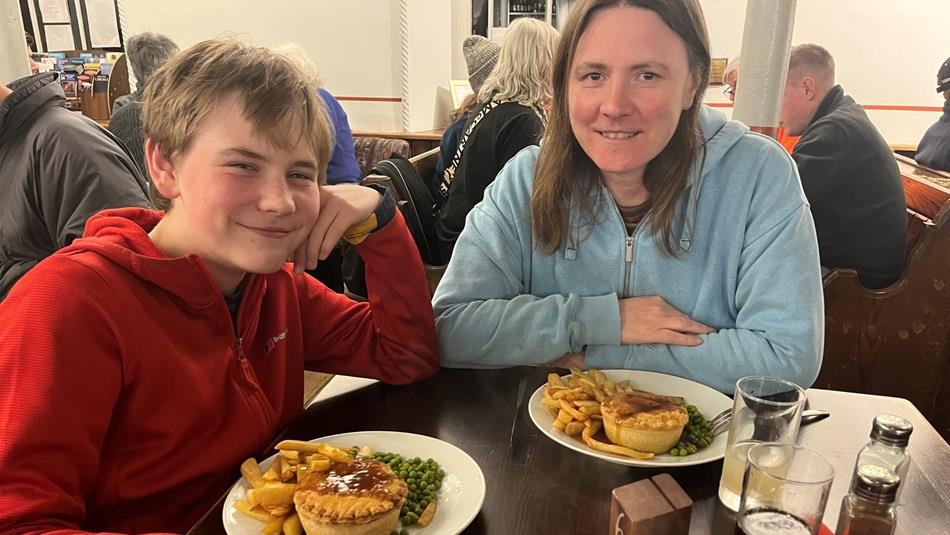 A woman and boy sitting at a table eating pie, chips and peas