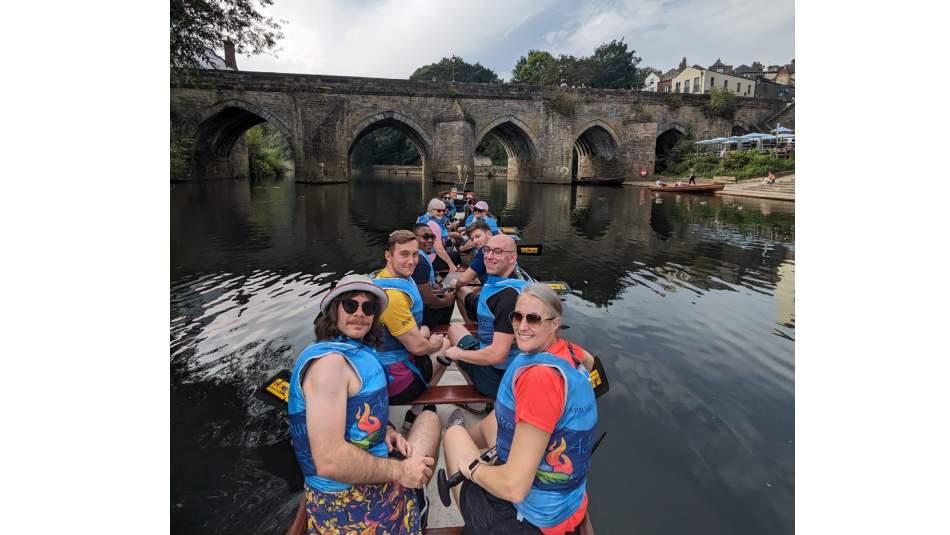 A group of people wearing life jackets paddling a dragon boat on a river, with an old stone arched bridge and riverside buildings in the background.