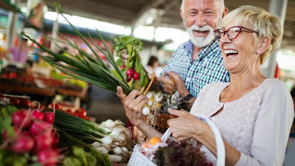 Woman and man smiling next to vegetable stall of market. Woman holding vegetables.