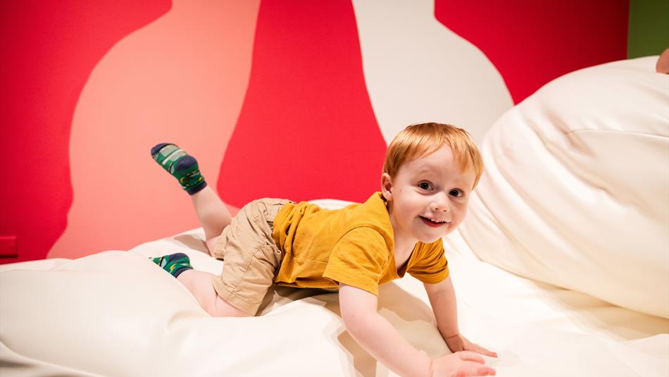 Photograph of a young child with red hair wearing a mustard yellow shirt and beige shorts, smiling while crawling on a large white cushioned surface.