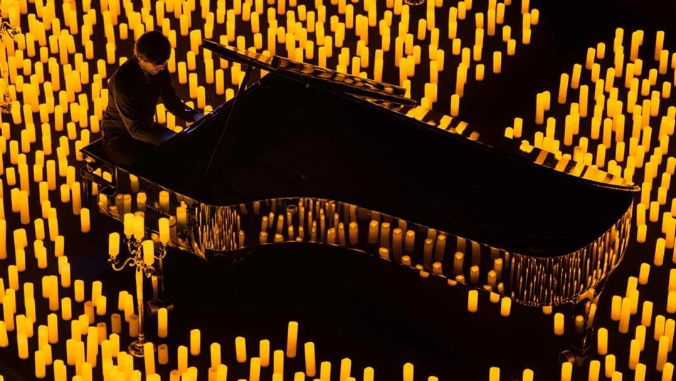 A man playing a grand piano surrounded by candles placed on the floor.