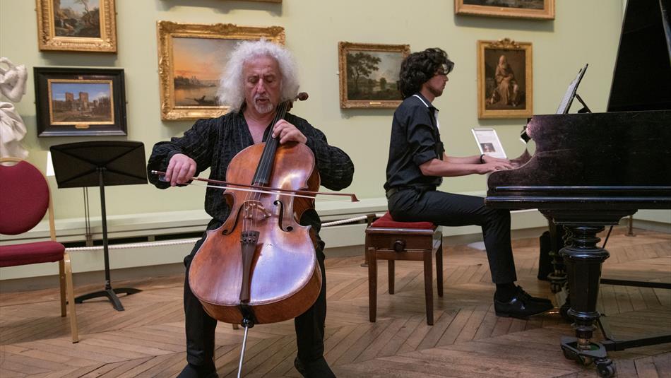 A man playing a cello and a man playing the piano room in the picture gallery.