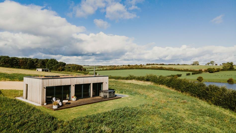 Modern wooden lodge with large glass windows and hot tub, set in a grassy hilltop overlooking green fields and a pond under a blue sky with scattered