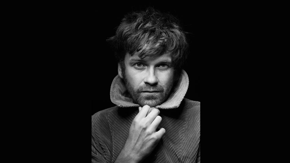 A black and white image of a man with brown hair and a beard looking straight at the camera.