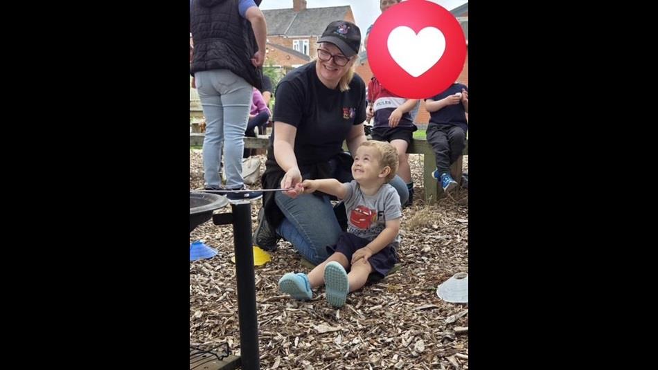 A child sitting on the ground toasting marshmallows, supervised by an adult - Forest School activities at St Johns Hall Meadowfield.