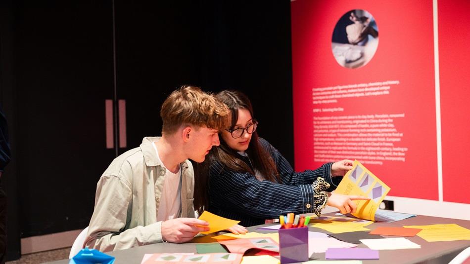 A young man with brown hair and a young woman with brown hair and glasses sat together making art in a bright pink space.