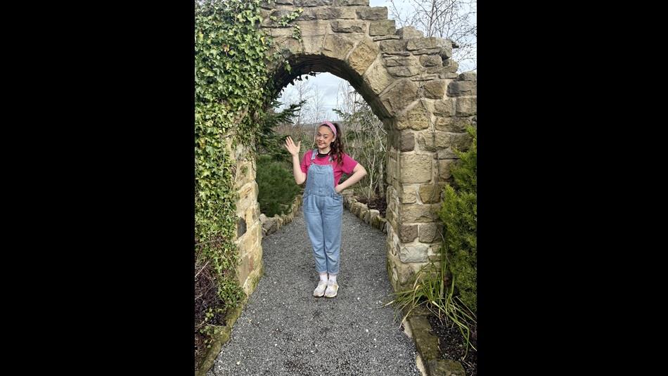 Miss Rachel waving at the camera, wearing dungarees and a pink t shirt.