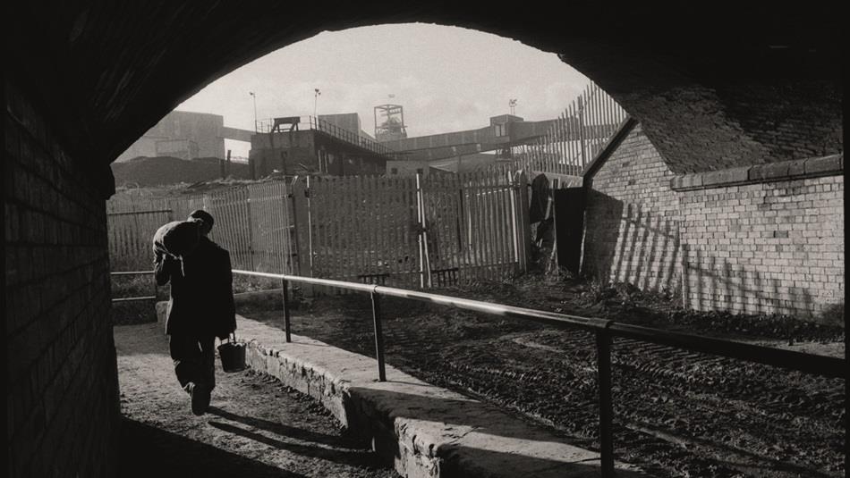 A photograph from the exhibition, a workman walking under railway bridge, carrying a bucket with a heavy sack over his shoulder
