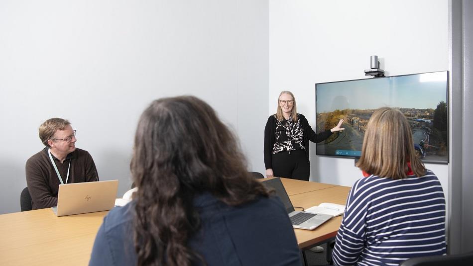 Meeting room with 4 people, three seated at table and one standing by display screen.