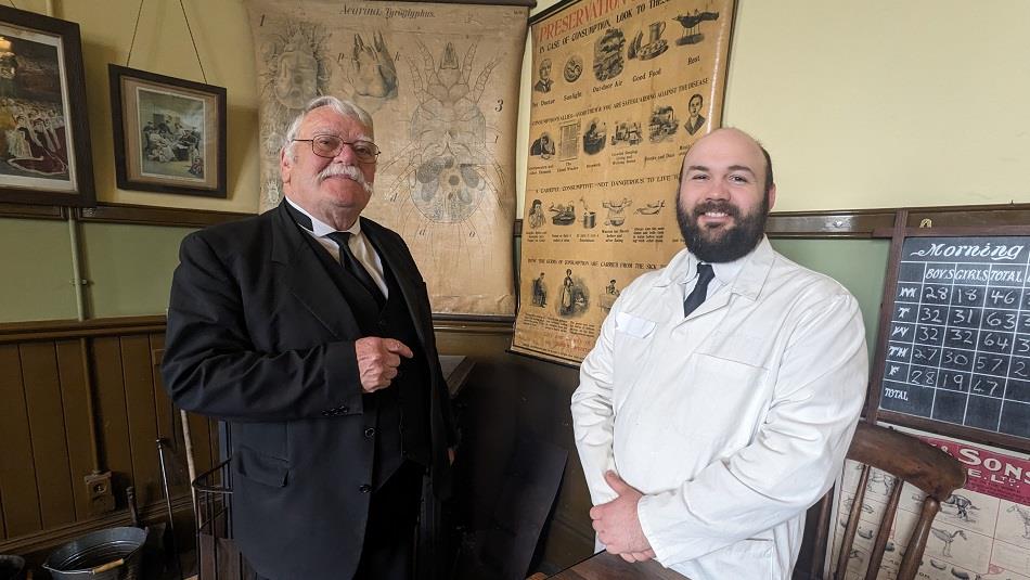 Two people in period clothing standing in the classroom at Beamish