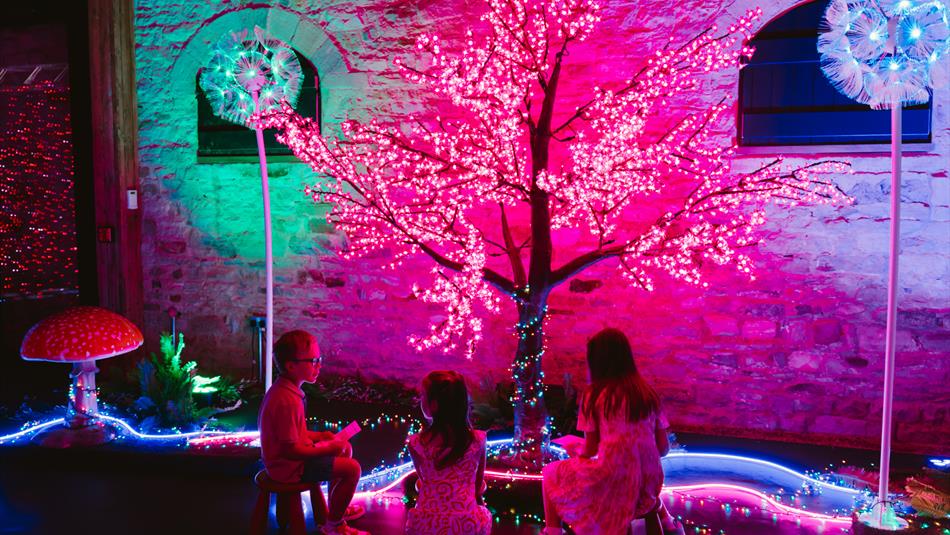 Children sitting under a tree with leaves made from pink fairy lights at 'The Hidden Garden: An Immersive Experience'  - Raby Castle, Park and Gardens