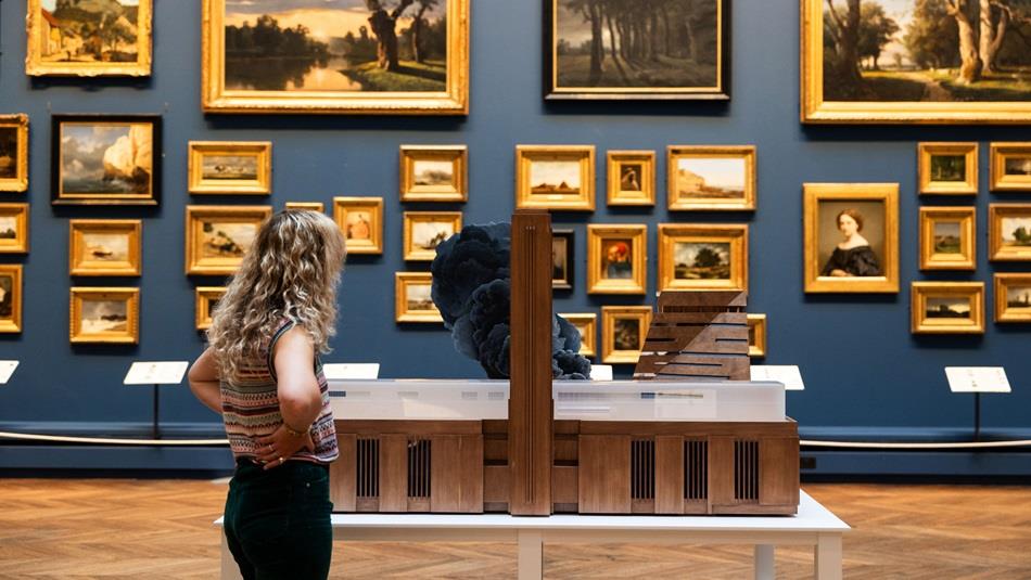 A woman with blonde curly hair looking at Tate Modern on Fire by Nathan Coley in a blue picture gallery with walls covered by paintings